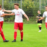 Fußballspieler in weißen Trikots feiern eine Toraktion, während zwei Mitspieler jubeln; im Hintergrund Schiedsrichter und Zuschauernähe auf dem Trainingsfeld.