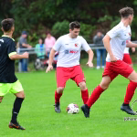 Drei Fußballspieler laufen auf dem Rasen um den Ball in einem Spielmoment; Hintergrundleute und Stadionabsperrungen verschwommen.