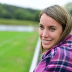 Porträt einer Frau mit Blick in die Ferne, warmes Lächeln; Hintergrund zeigt einen grünen See und Wiesenlandschaft.