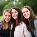 Drei lächelnde Frauen posieren eng nebeneinander im Freien, Hintergrund unscharf, Nahaufnahme für einen freundlichen Gruppenmoment.