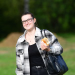 Lächelnde Person im Freien, hält eine süße Teilchenrolle in der Hand; trägt Jacke und Handtasche, Hintergrund grün unscharf.