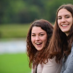 Zwei junge Frauen posieren im Freien, lachen in die Kamera; unscharfer Hintergrund für einen freundlichen Porträt-Eindruck.