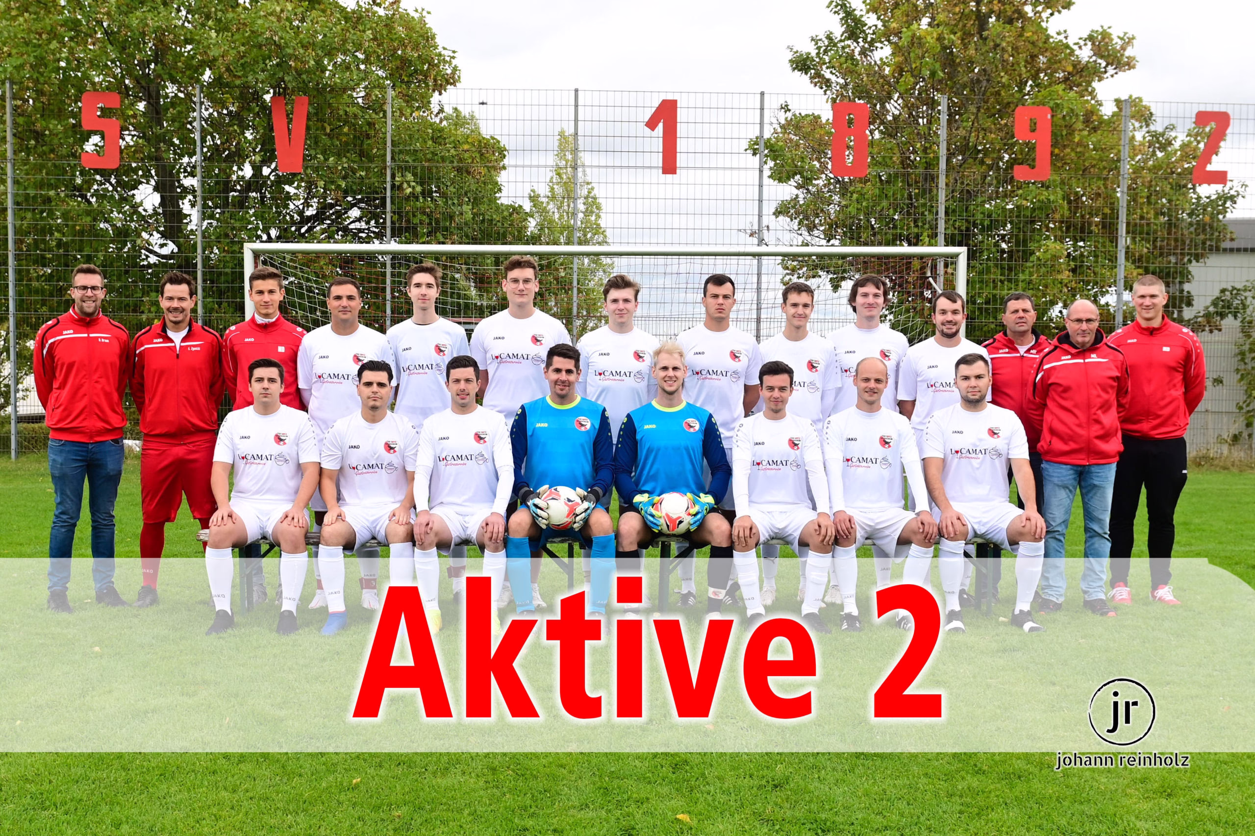Fußballmannschaft im Gruppenfoto vor Tor und Stacheldrahtzaun, darüber rote Zahlen 5 V 1 8 9 2 sowie Titel „Aktive 2“.